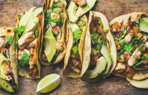 six soft tacos on a wooden tabletop with avocado and sliced limes