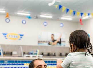 a dad reaching for his toddler daughter who is sitting on the edge of a pool, ready to get in and swim with him