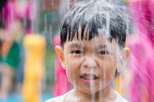 an Asian toddler boy playing at a splash pad while water falls on his head