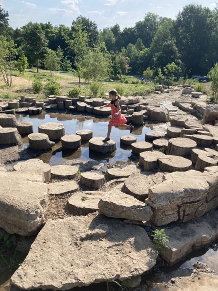 a girl jumping from stump to stump across a stream at Forest Park and the Nature Playscape feature