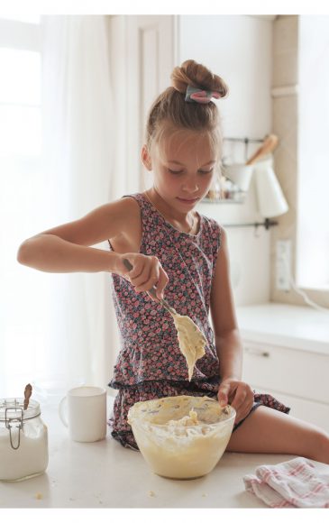 a girl sitting on the counter as she holds a spoon, dripping batter, over a bowl