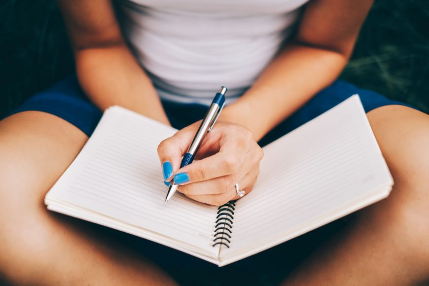 a close up of a woman, with blue nail polish, holding a pen as she is journaling in a notebook