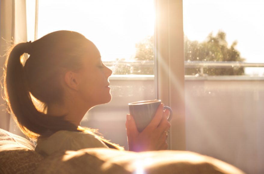 a woman sitting on a couch as the sun rises through the window, drinking coffee
