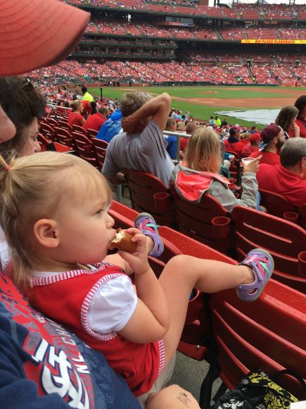 a toddler girl sitting on her dad’s lap as they watch Cardinals baseball