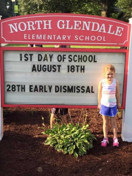 an incoming kindergarten girl standing next to the North Glendale elementary school sign, as she gets ready for the first day of school