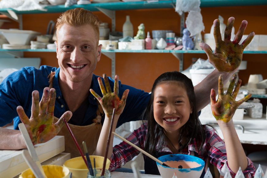 a girl and a man painting pottery, with their hands raised and covered with paint