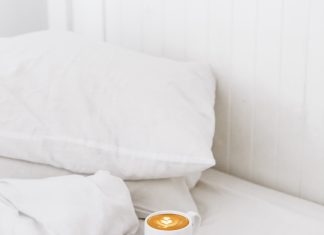 a bed of white linens and pillows, with a mug of coffee, a book, and glasses set on the comforter to represent self-care