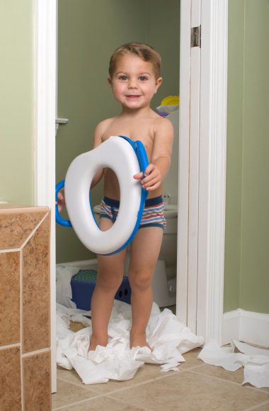 a toddler boy standing in a bathroom doorway, surrounded by unspooled toilet paper, and holding a potty seat