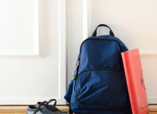 a navy blue backpack sits by a closed door, with a red notebook leaning on it and a pair of shoes nearby getting ready for back-to-school