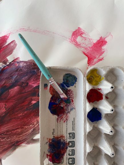 An egg carton with yellow, red, and blue paint in three compartments is shown. The lid of the egg carton has paint that has been mixed on it. A paintbrush rests in the tray. Beneath the egg carton is a white piece of paper with red, blue, and yellow brushstrokes. 