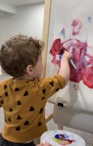 A toddler wearing a yellow and black patterned long sleeved t-shirt is shown painting on an easel with a paint brush. Red, pink, and blue brushstrokes are seen on the white paper in circular motions.