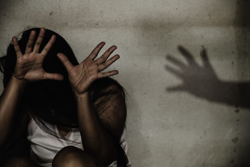 a woman huddling in a corner with her arms raised as the shadow of a man’s hand comes at her