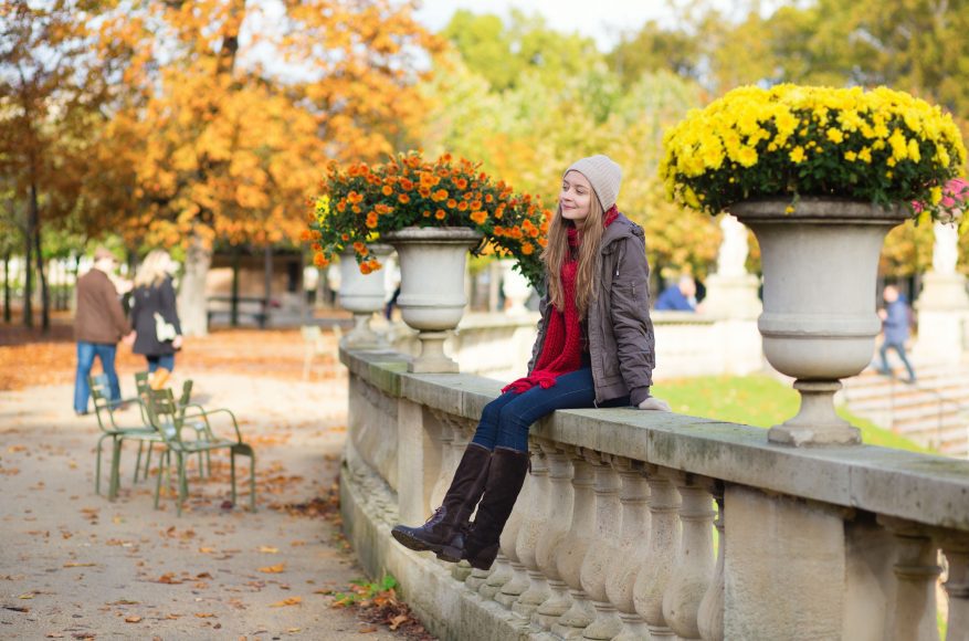 a woman wearing fall fashion pieces such as a beanie and tall boots as she sits on a wall on a fall day