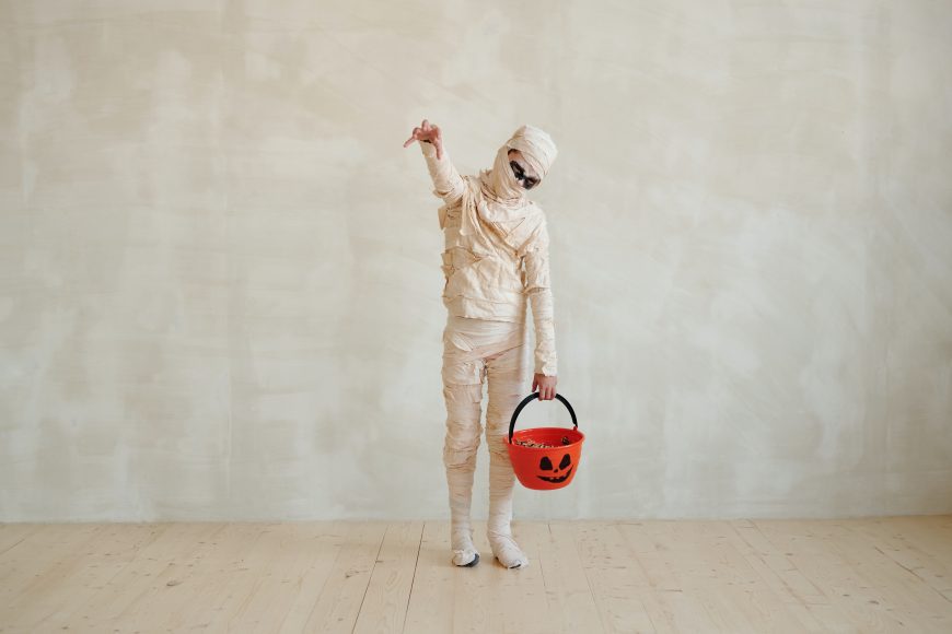 a child in a mummy costume with a pumpkin trick or treat bucket