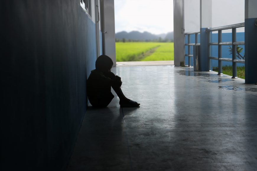 a teen boy sitting in a dark hallway with his hands wrapped around his knees