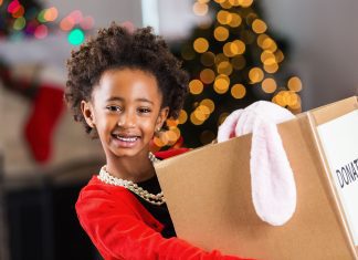 a young African American girl in a bright red sweater holding a box of toy donations as she stands in front of a Christmas tree