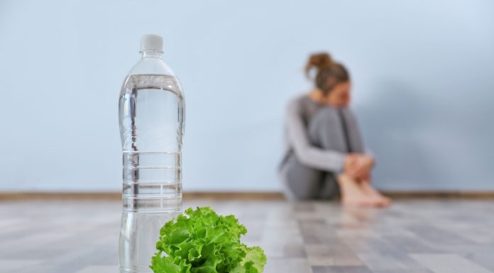 a woman in the background, sitting on the ground as she hugs her knees to her and a bottle of water and piece of lettuce sit in the foreground