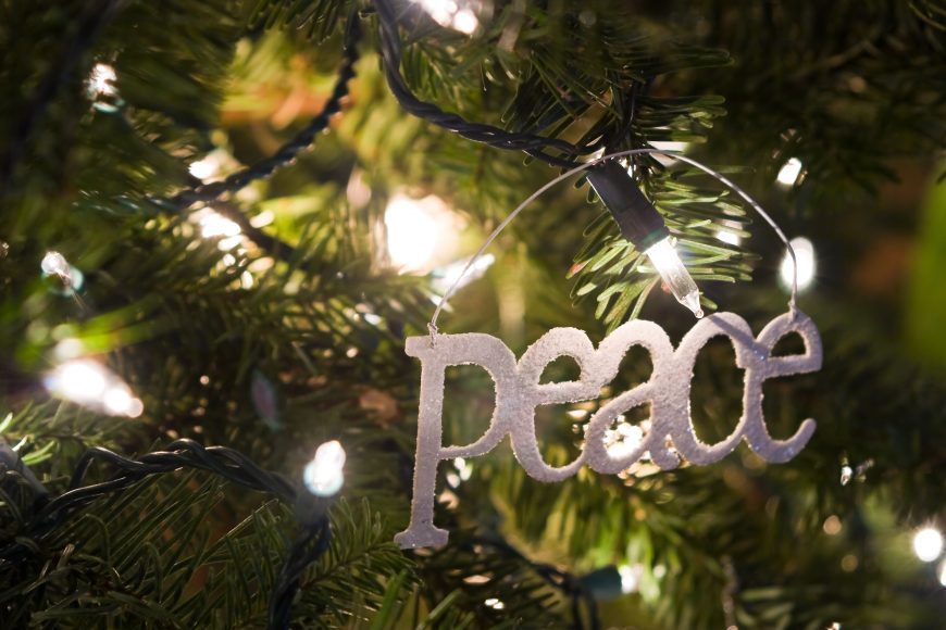 a silver ornament with the word, “peace” hanging on an evergreen branch