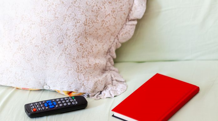 a red, hard cover book on a bed next to a pillow and a TV remote