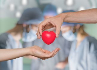 an operating room out of focus in the background with a hand in the foreground, handing a plastic heart to an open palm