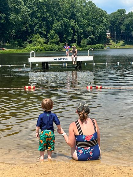 a mom and her toddler son sitting at the edge of a lake as they watch kids jump in from a platform