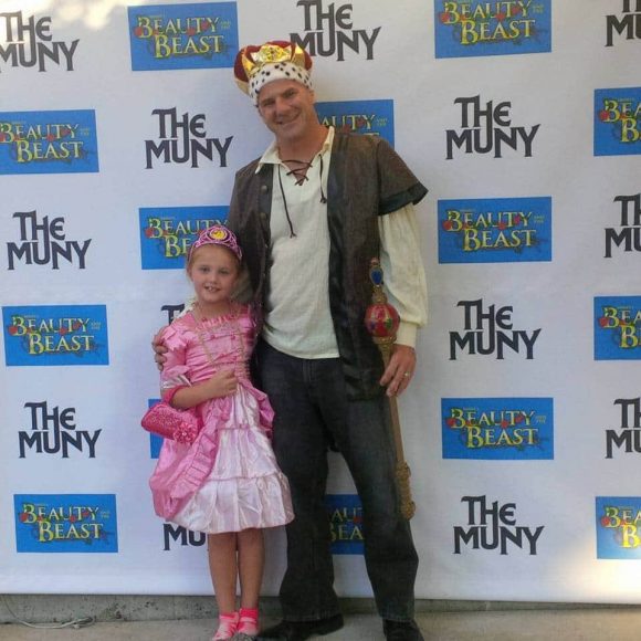 A dad dressed as a king, and his daughter dressed as a princes in front of the Beauty and the Beast backdrop at The Muny in St. Louis