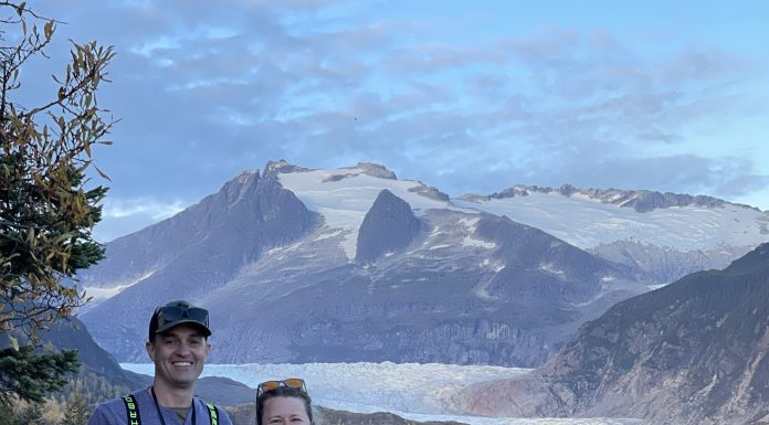 a husband and wife take a trip together as they stand in front of a snowy mountain by a river