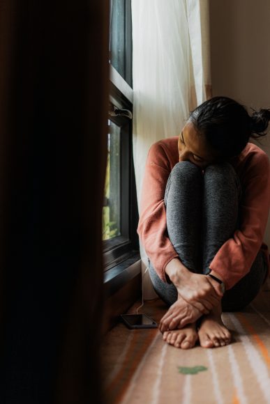 a woman sitting on the floor, arms wrapped around her folded up legs, staring out the window as she battles anxiety and depression