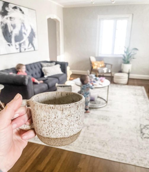 a mom holding up a cup of coffee with her child playing on the couch in the background as it gets colder outside