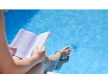 a photo from above of a woman sitting with her feet in the pool as she reads a book