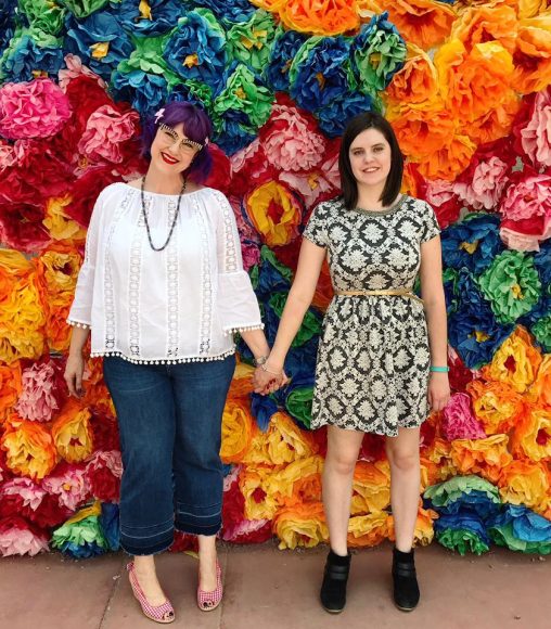 a mom and her adult daughter pose, holding hands in front of a wall of flowers