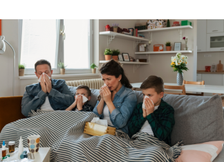 a mom, dad, and their two kids all under a blanket on the couch. They all have tissues to indicate they are sick with viruses