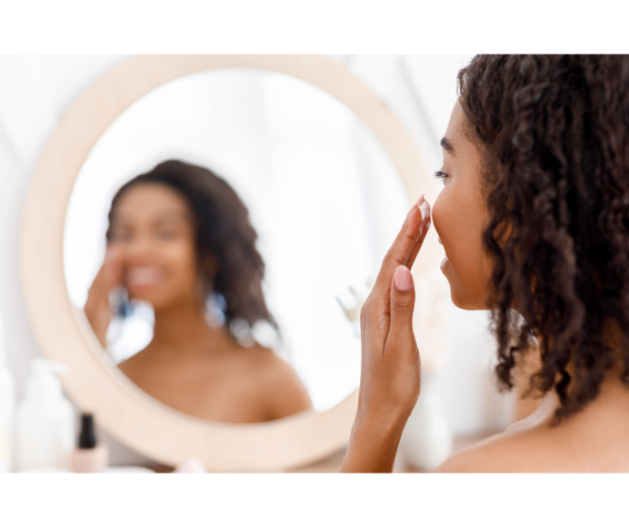 an African-American woman applying lotion to her face as she looks in a mirror 