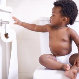 an African American toddler boy sitting on a closed toilet seat as he plays with the toilet paper roll.