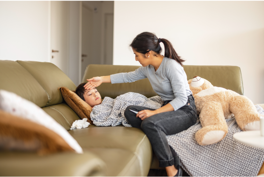 a child on a couch under a blanket as his mom sits next to him with her hand on his forehead and a thermometer in her other hand