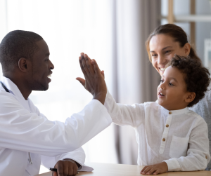 an African American pediatrician in a white lab coat with a stethoscope around his neck, giving a high five to a child who sits on his mom's lap