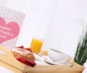 a wooden tray with a cinnamon roll, coffee, and orange juice next to a card with a pink heart that says, "Happy Mother's Day"