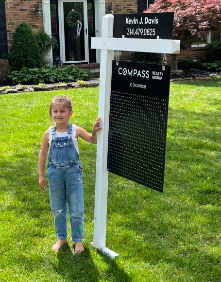 as moving day approaches, a young girl stands next to a for sale sign in her yard