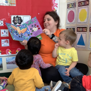 a preschool teacher reading a book to three children sitting near her on the carpet 