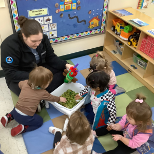 a group of preschool children sitting on a carpet with their Kiddie Academy teacher