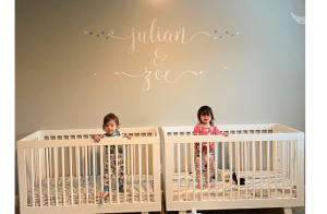 2 toddlers standing up inside their white wood cribs looking at the camera. The names Julian and Zoe are written on the wall behind them. 