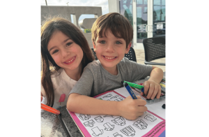 little girl with dark brown hair and little boy with light brown hair who is doing an activity book both smiling at the camera.