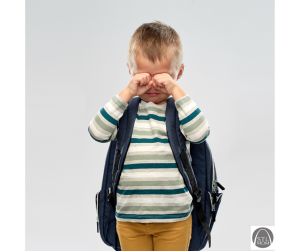 a young boy wearing a backpack, wiping tears from his eyes as he has a meltdown after school