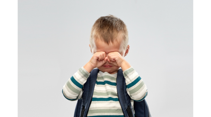 a young boy wearing a backpack, wiping tears from his eyes as he has a meltdown after school