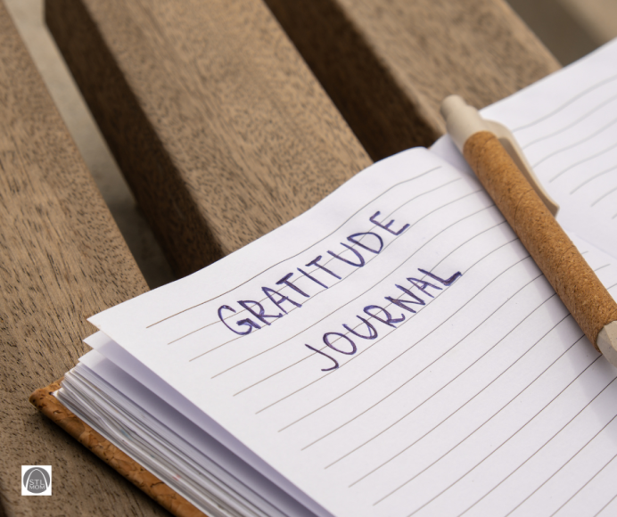 a journal open on a wooden bench. The journal has the words "gratitude journal" written on a lined page, and there is a pen on the page