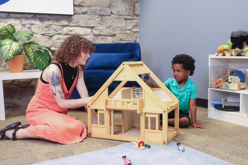 a therapist sitting on the floor with a child as they play with a dollhouse at Bud to Bloom Play Therapy