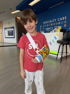 young boy in a red t-shirt smiling with his left arm in a blue cast and multi-colored sling in the lobby of a doctors office