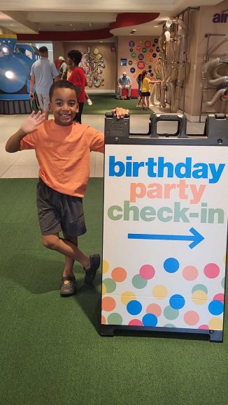 a young African-American boy in an orange shirt and gray shorts standing in front of a sign that says, "birthday party check-in" at the Magic House in St. Louis