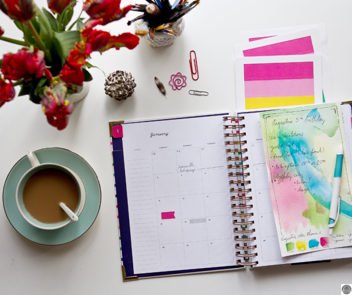 a planner open on a table next to a cup of coffee and a vase of flowers