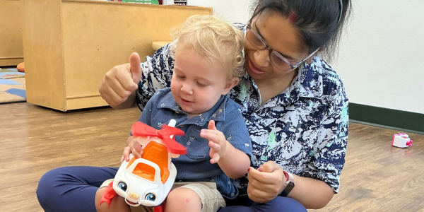 a teacher fostering connection with a child who is sitting in her lap, playing with a helicopter toy